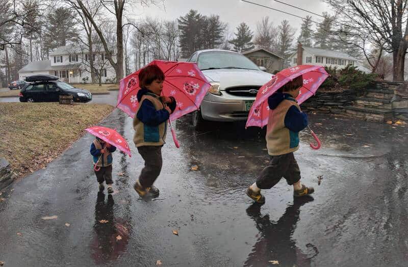 Three children walking with pink umbrellas on a rainy day in a suburban neighborhood.