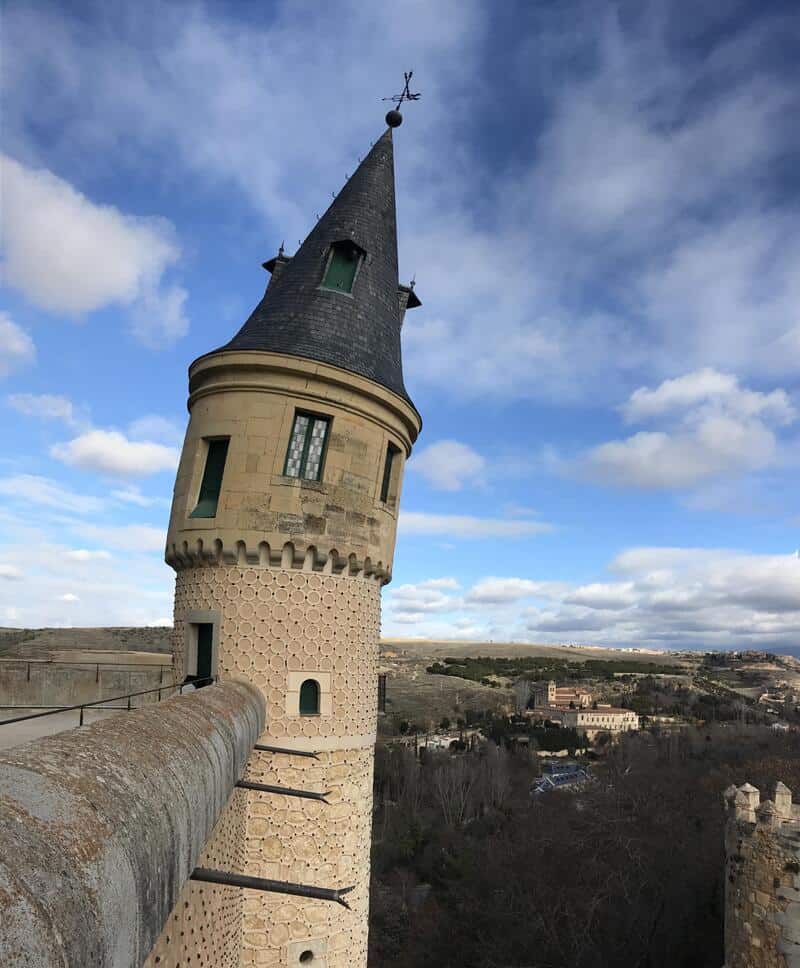 Tall castle tower with a pointed roof and a weather vane, overlooking a landscape.