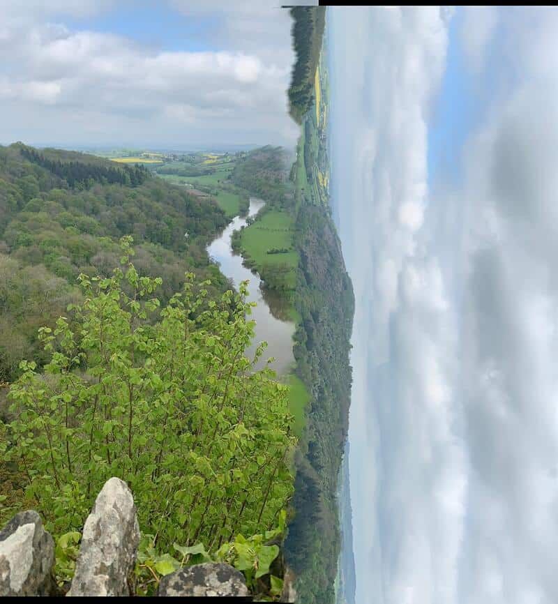 Panoramic view of a river winding through green hills and forests under cloudy sky.