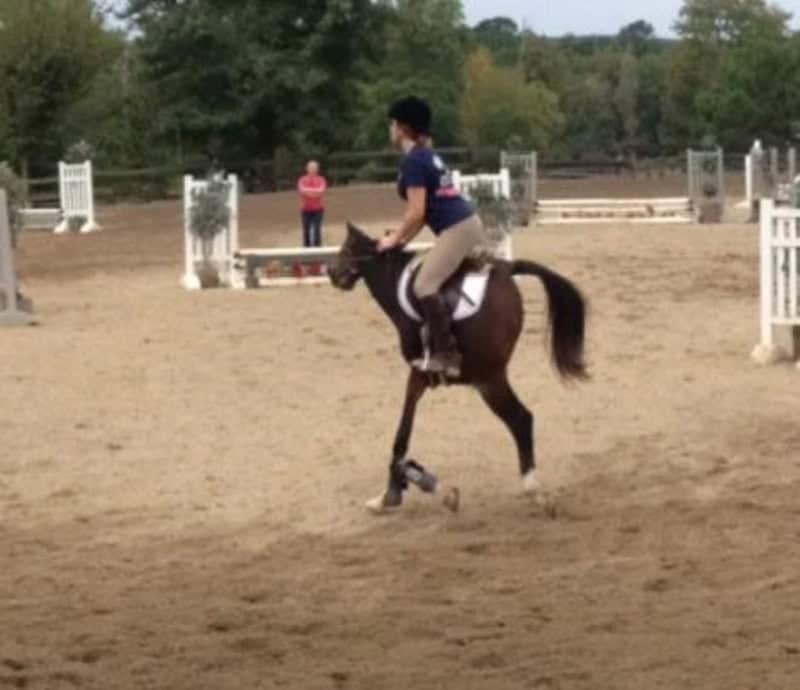 Person riding a horse in an outdoor arena with a woman in pink watching.