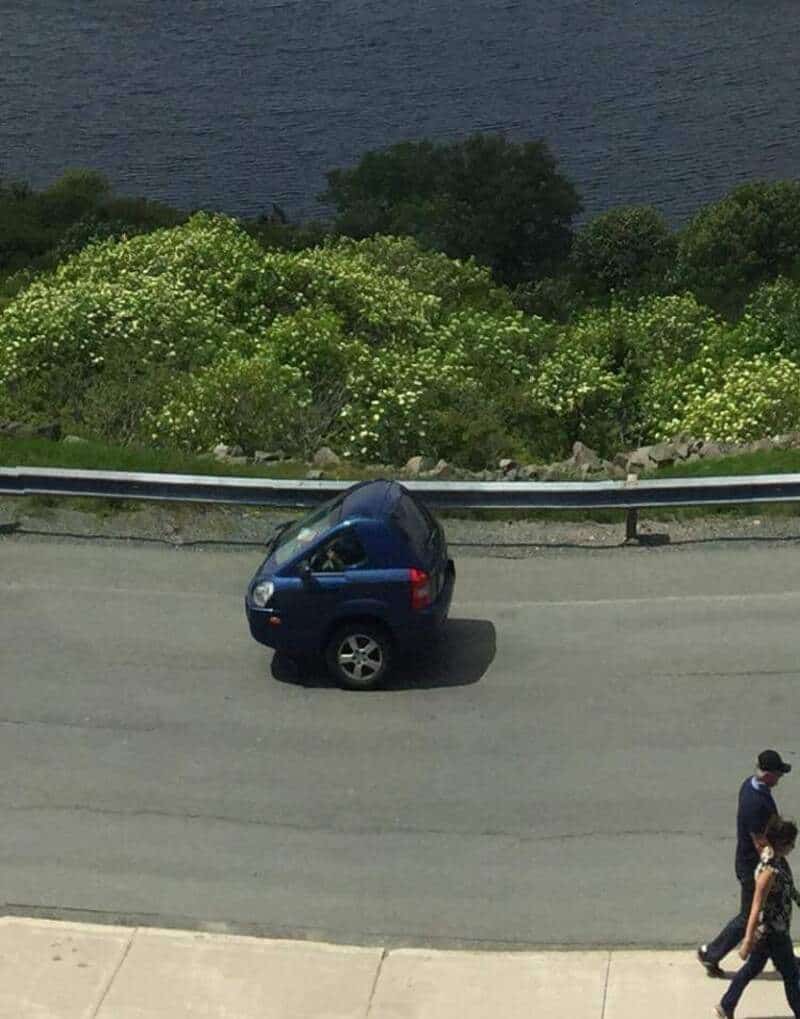Small blue car on a curved road with water and greenery in the background.