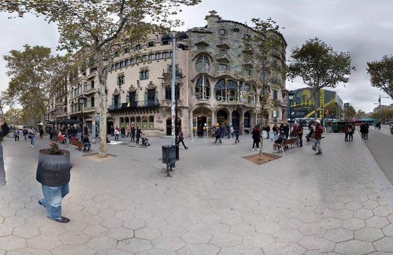 Panoramic view of a busy city square with a unique, curved building and people walking around.