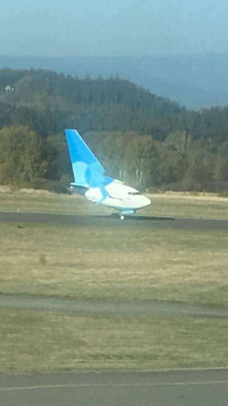 Airplane with a blue tail on a runway, with a forest and mountains in the background.