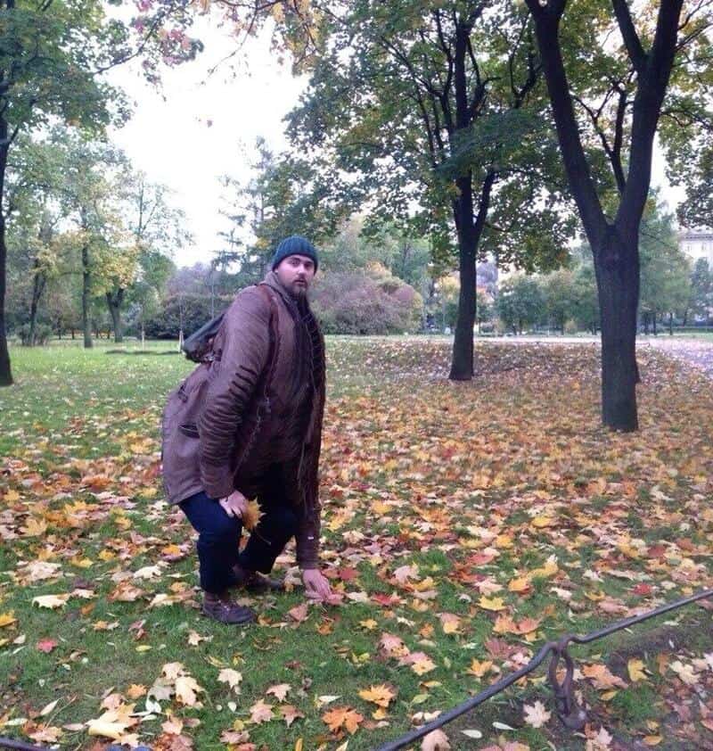 Man kneeling on the ground in a park with fallen leaves, trees, and a cloudy sky.