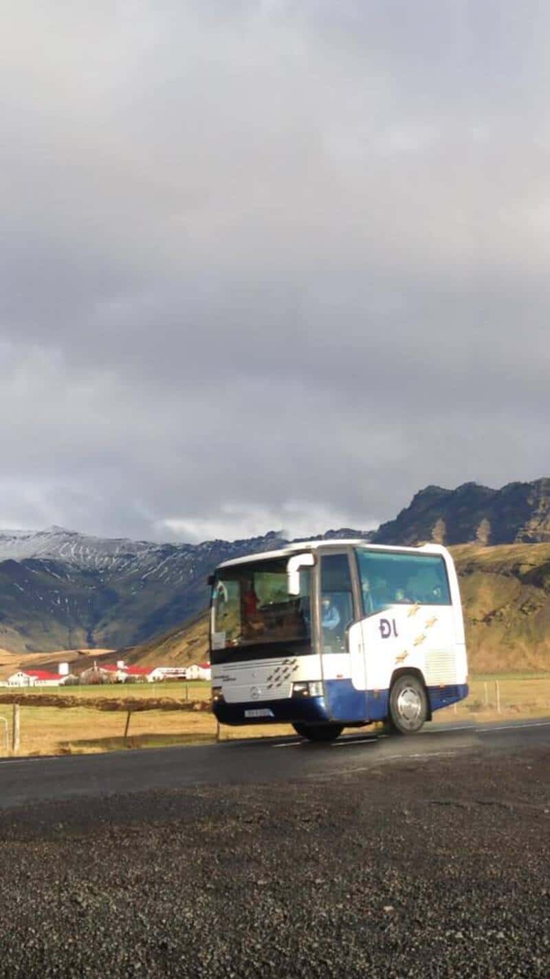 A small bus on a rural road with mountains and cloudy sky in the background.