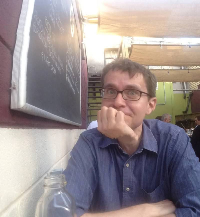Man with glasses in a blue shirt sitting at a table in a cafe, resting his chin on his hand.