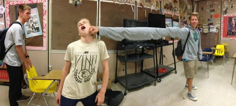 Three boys in a classroom, one with a long paper tube, two reacting humorously.