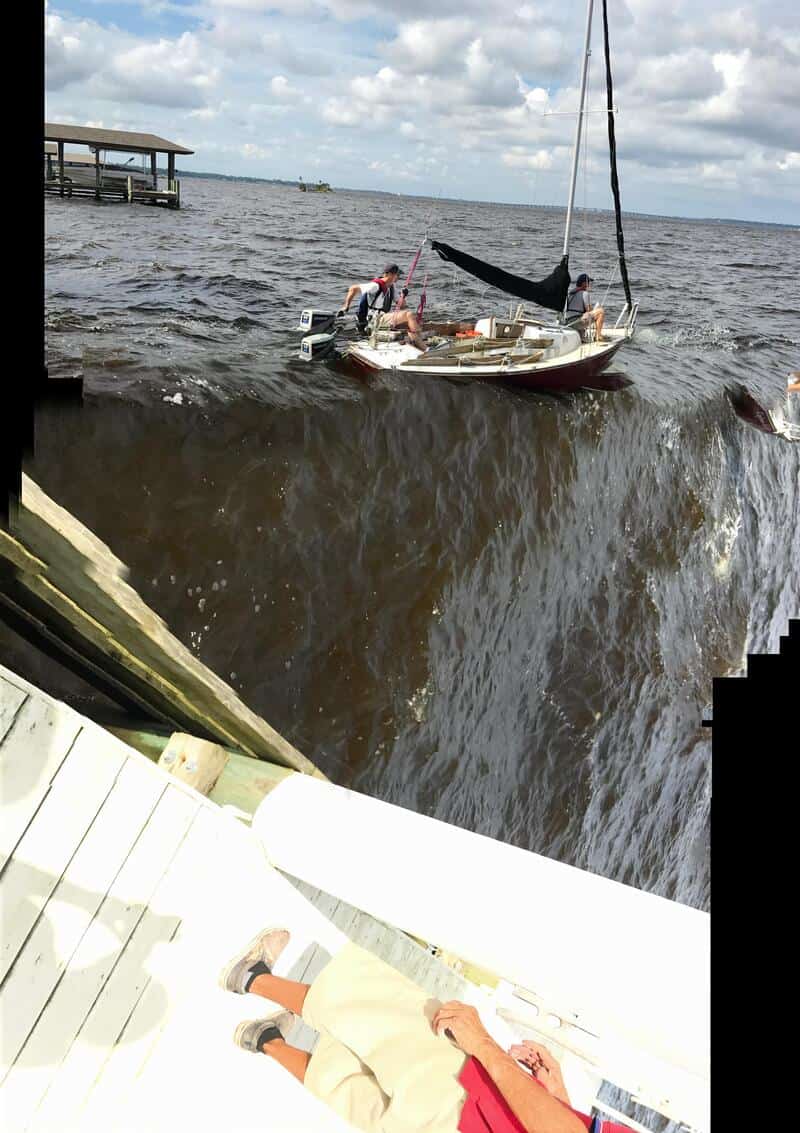 Sailboat capsized in choppy water with two people on it, near a dock.
