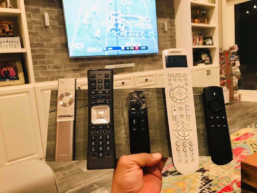 Five remote controls held in a hand in front of a TV and a living room.