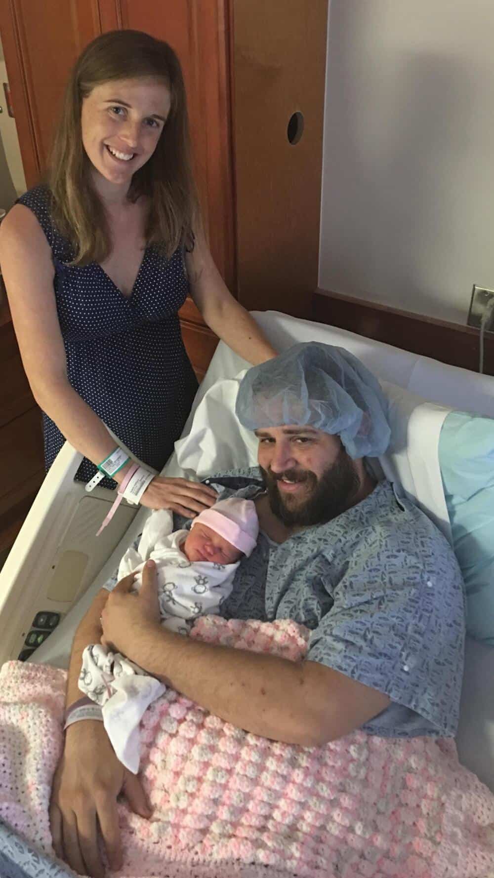 Happy woman with hospital wristbands, man in hospital gown with cap, holding newborn baby.