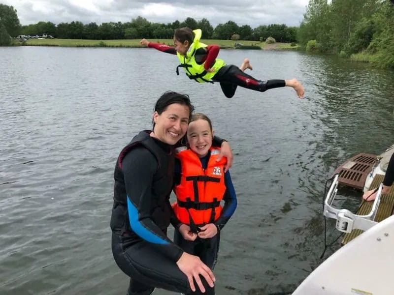 A woman and girl in life jackets by a lake, with a boy flying in the air above them.
