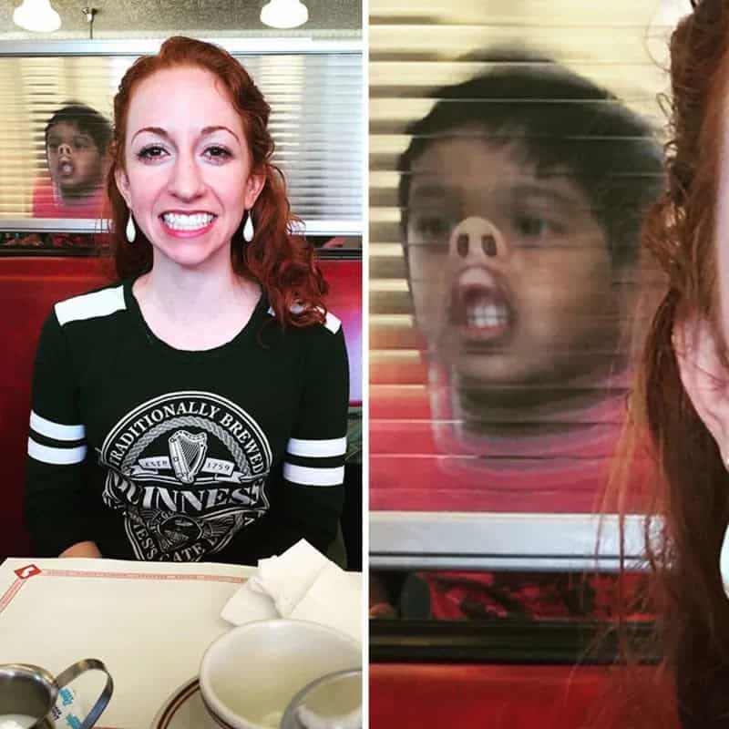 Woman smiling at camera with a child's face pressed against window behind her.