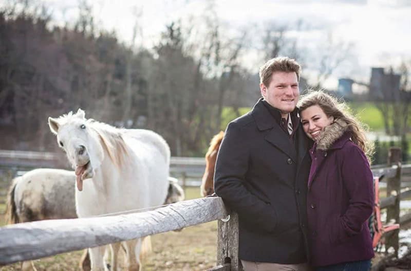 Couple smiling near horses at a farm, with one horse photobombing.