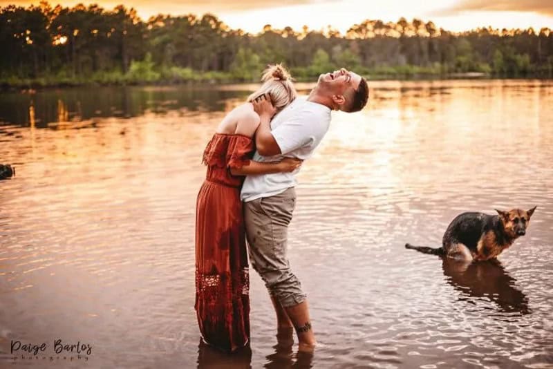 Couple hugging in water with a dog nearby at sunset.