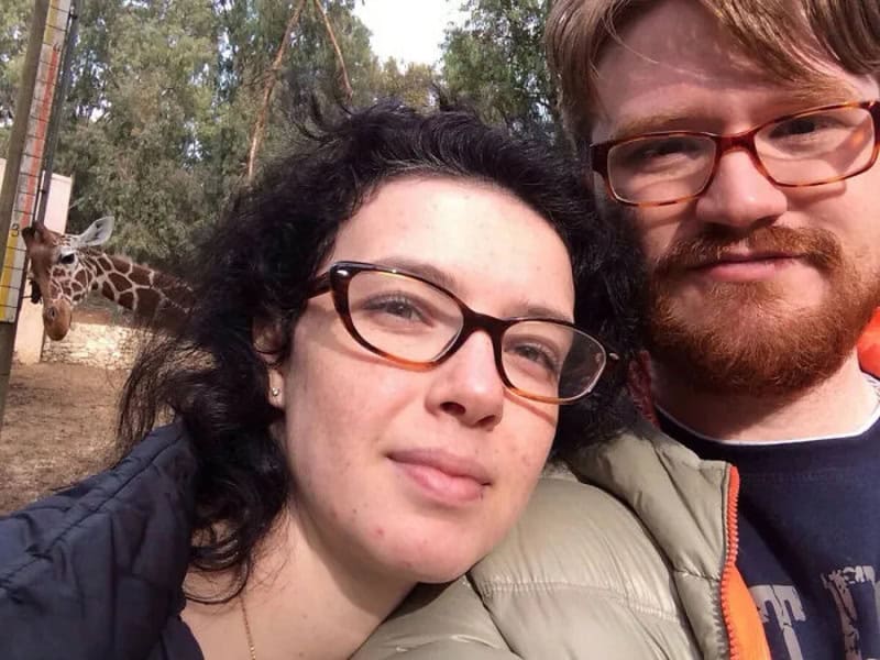 A woman and man taking a selfie outdoors with a giraffe in the background.