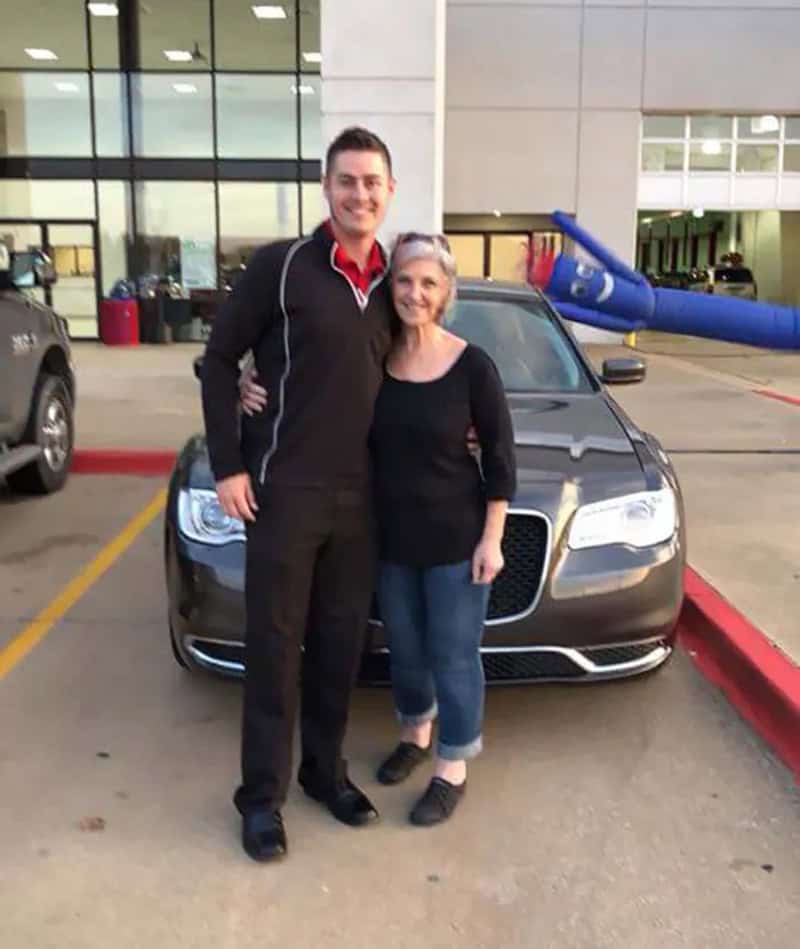 A young man and an older woman stand in front of a black car at an airport.