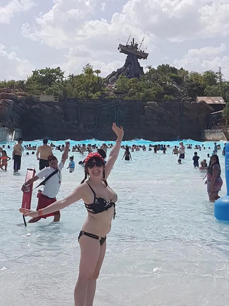 Woman in black bikini waving at water park with pirate ship in background.