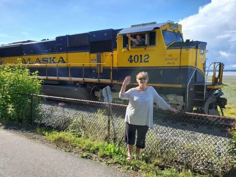 Woman waving in front of a yellow Alaska train with a person in the cab.