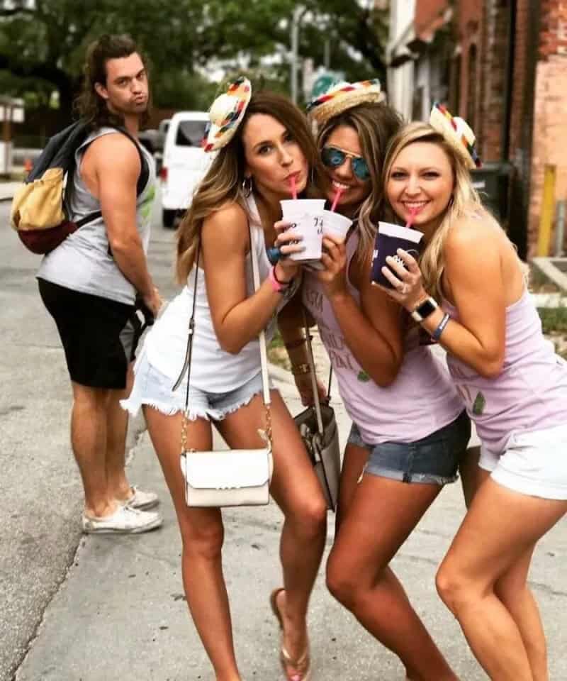 Three women in summer clothes with party hats and drinks, one man in background on street.