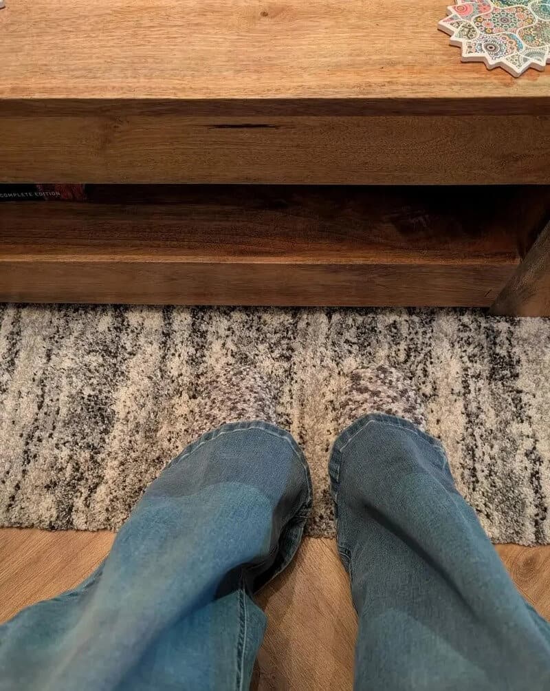 View of person's legs in jeans, carpet, and wooden furniture with decorative items on top.