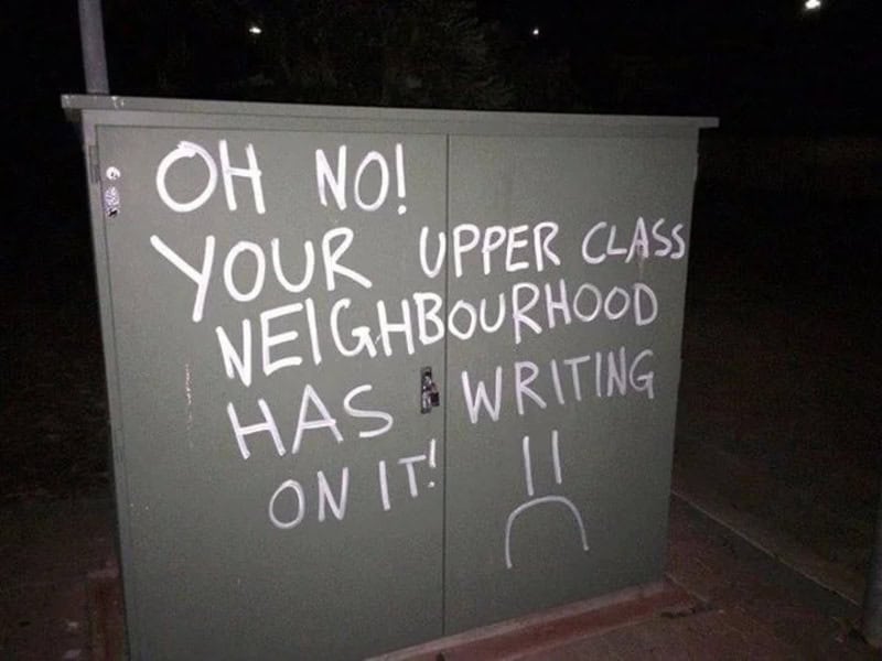 Graffiti on a grey electrical box says 'OH NO! YOUR NEIGHBOUR HAS WRITING ON IT!' with a sad face.