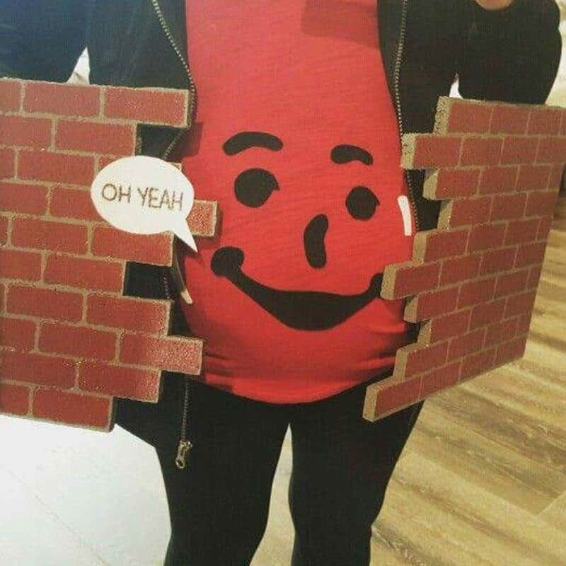 Pregnant woman in a red shirt with a smiley face, holding brick wall cutouts, speech bubble says 'OH.