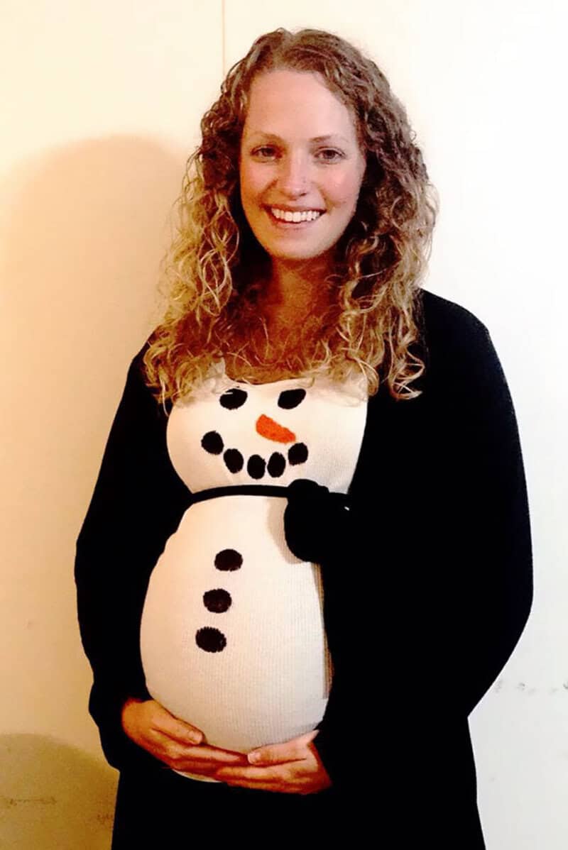 Pregnant woman in a snowman costume with a carrot nose and coal buttons.