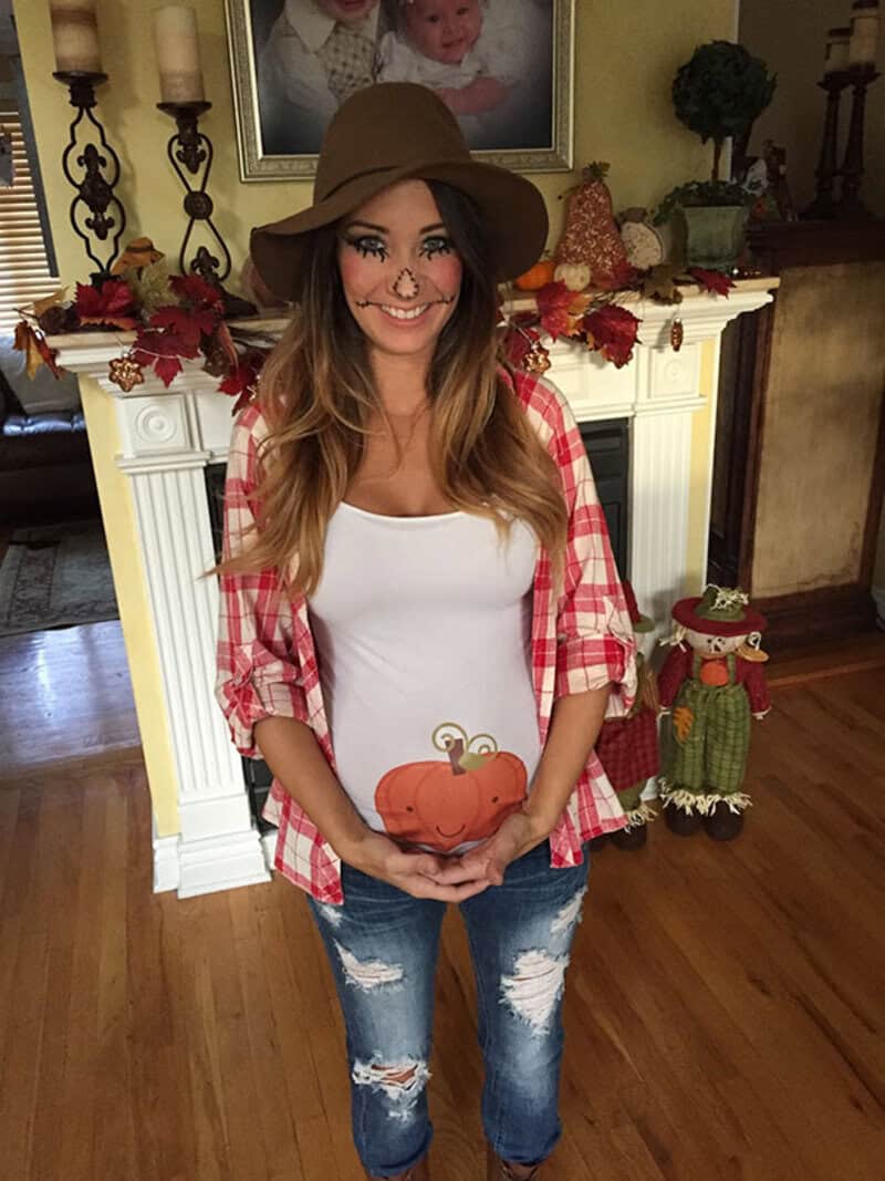 Pregnant woman in fall-themed outfit holding pumpkin, smiling in living room.