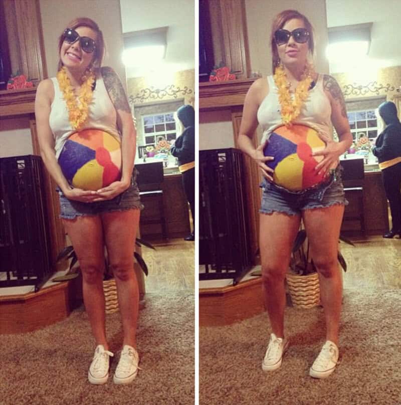 Pregnant woman in sunglasses, lei, shorts, holding beach ball, in kitchen.