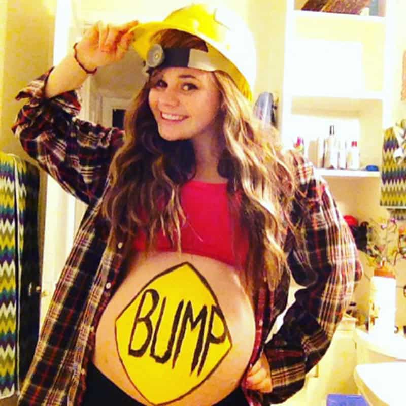 Pregnant woman in firefighter costume with 'BUMP' sign on belly, wearing yellow helmet.