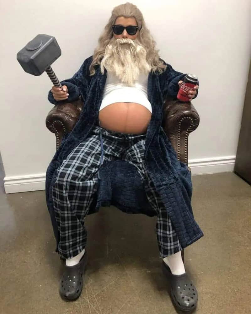 Pregnant man dressed as Thor with hammer and soda, sitting on a chair.