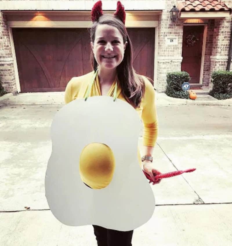 Woman in yellow with devil horns holding fried egg costume, smiling outside.