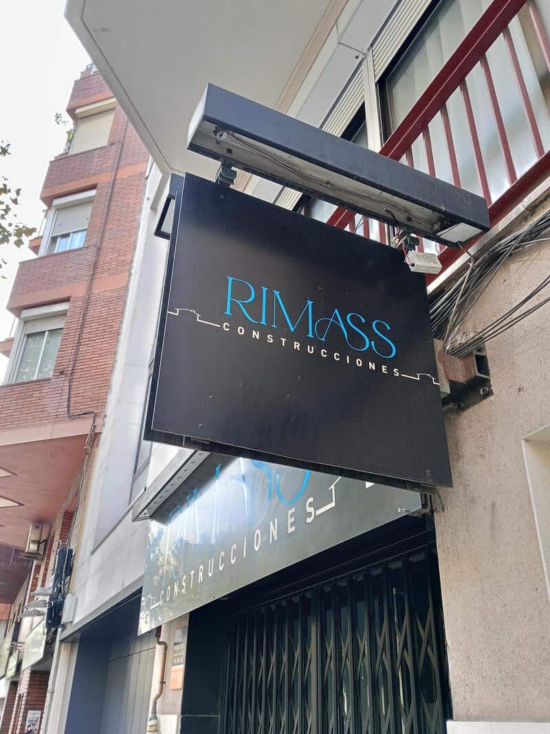 RIMASS Construcciones sign hanging on building exterior with blue text on black background.