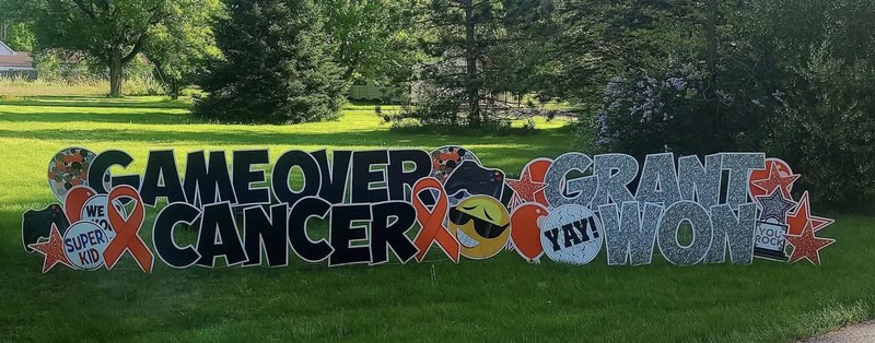 Sign reads 'GAME OVER CANCER' with ribbon icons, celebratory symbols and messages like 'WE WON' in a.