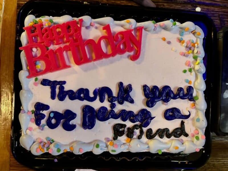 Happy Birthday cake with pink 'Happy Birthday' topper and blue writing 'Thank you for being a friend.