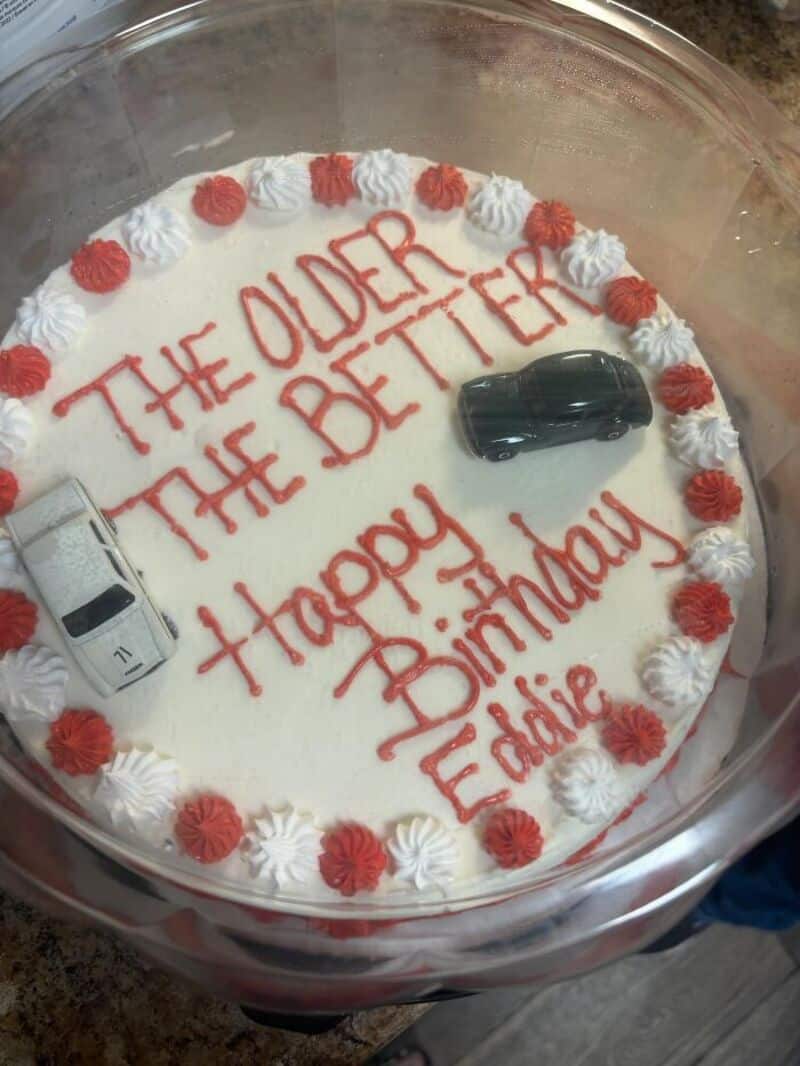 The older the better happy birthday Eddie cake with white frosting red cursive writing and toy cars.