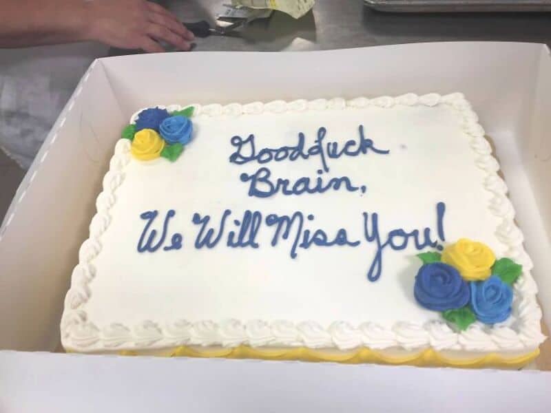 Hilarious cake with blue icing text: "Goodluck Brain, We Will Miss You!" and decorative yellow, blue.