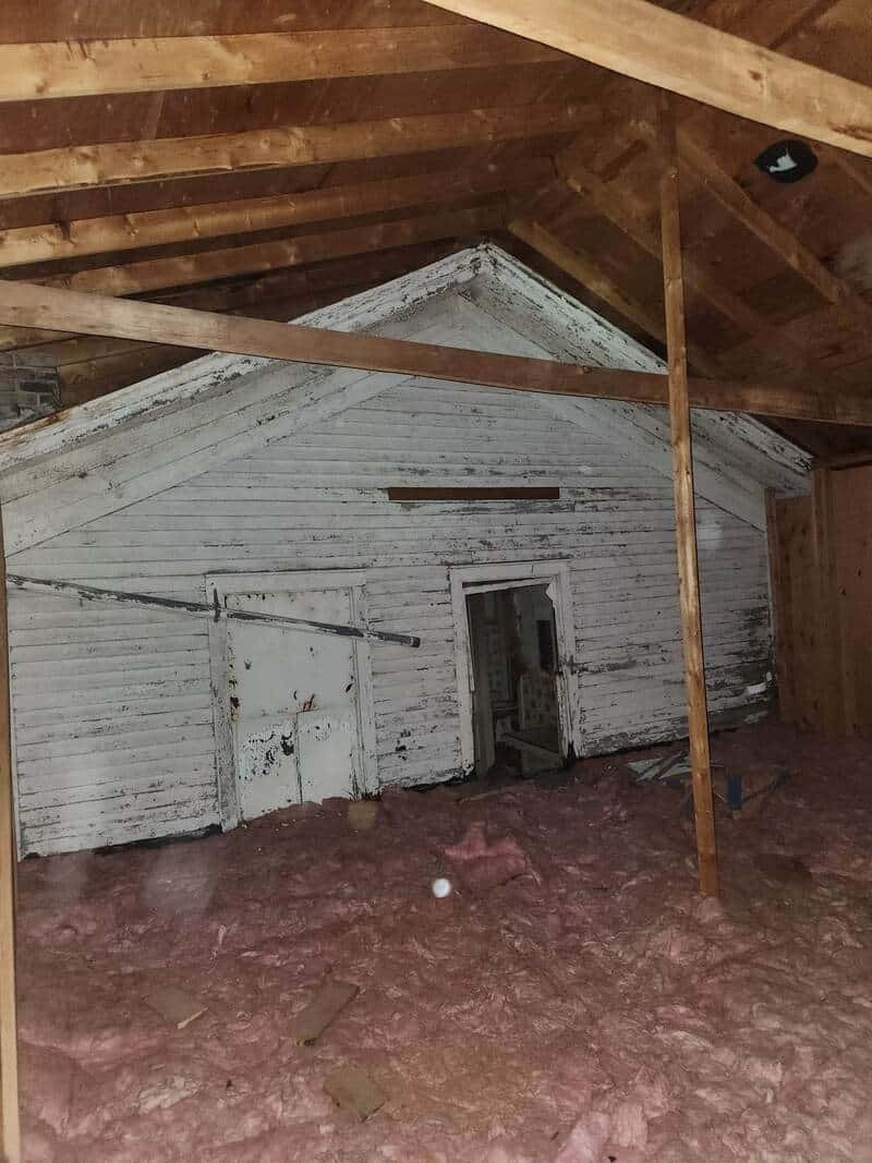 Attic space with exposed wooden beams, white painted wall, and pink insulation on the floor.