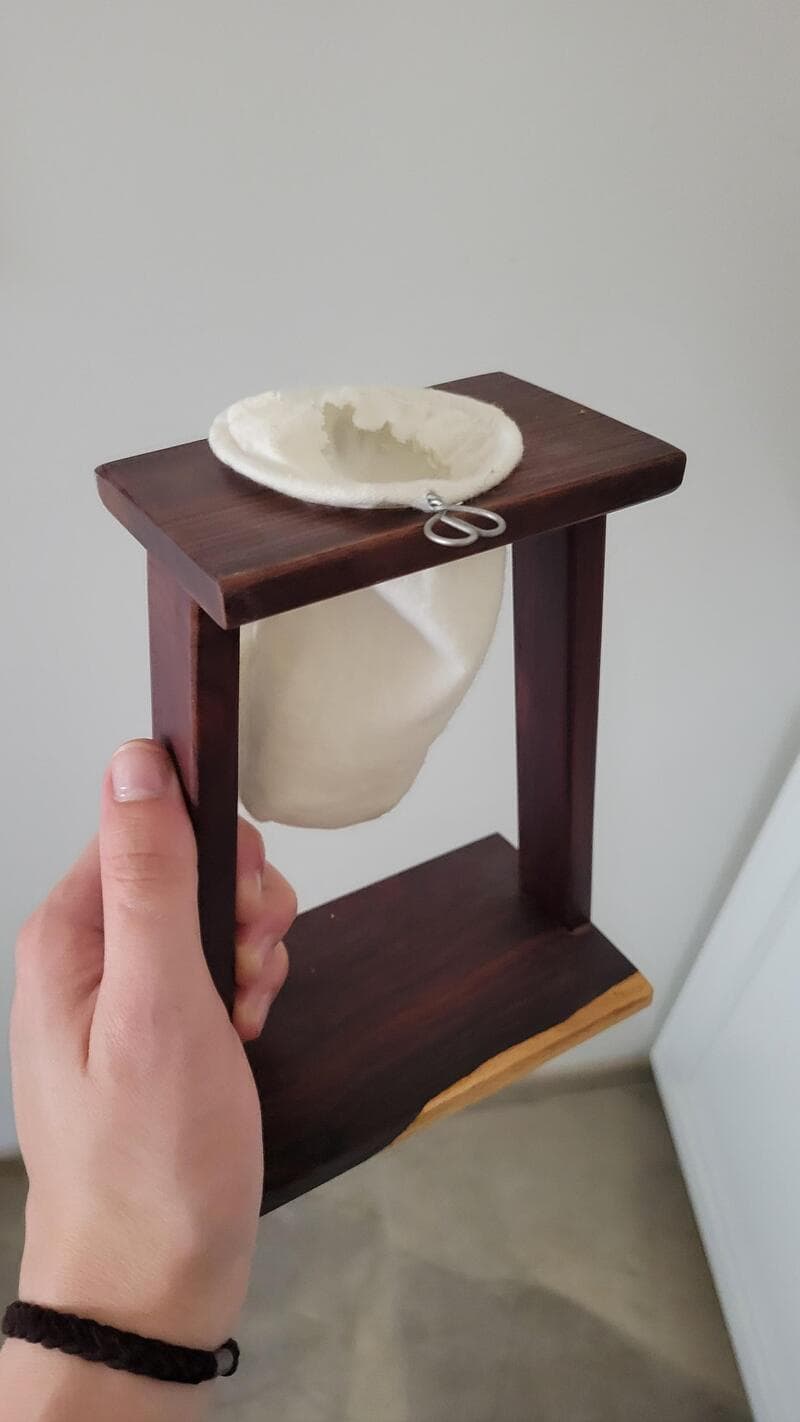 A hand holding a wooden stand with a white, hollow, egg-shaped object on top.