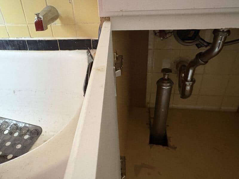 Underneath a kitchen sink showing pipes and a cabinet with an open door.