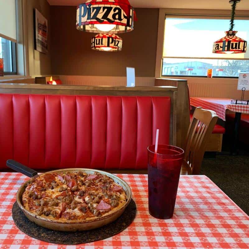 Pizza on a round pan with pepperoni and bacon slices on a red-and-white checkered table in a dine-in.