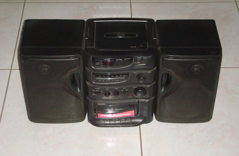Compact stereo system with central unit and two side speakers on a tiled floor.