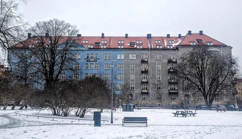 Snow-covered park with benches and trees in front of a blue and gray apartment building.