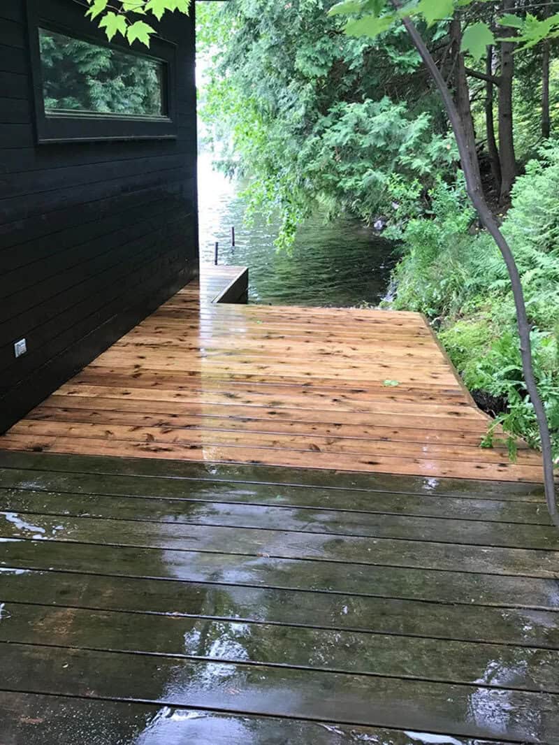 Clean and wet wooden deck beside a black house with green trees and water.