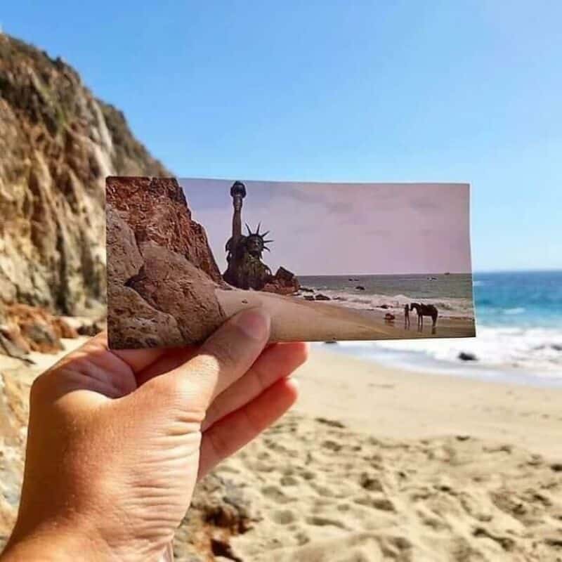 Hand holding a photo of the Statue of Liberty on a beach with rocks in the foreground and ocean in a.