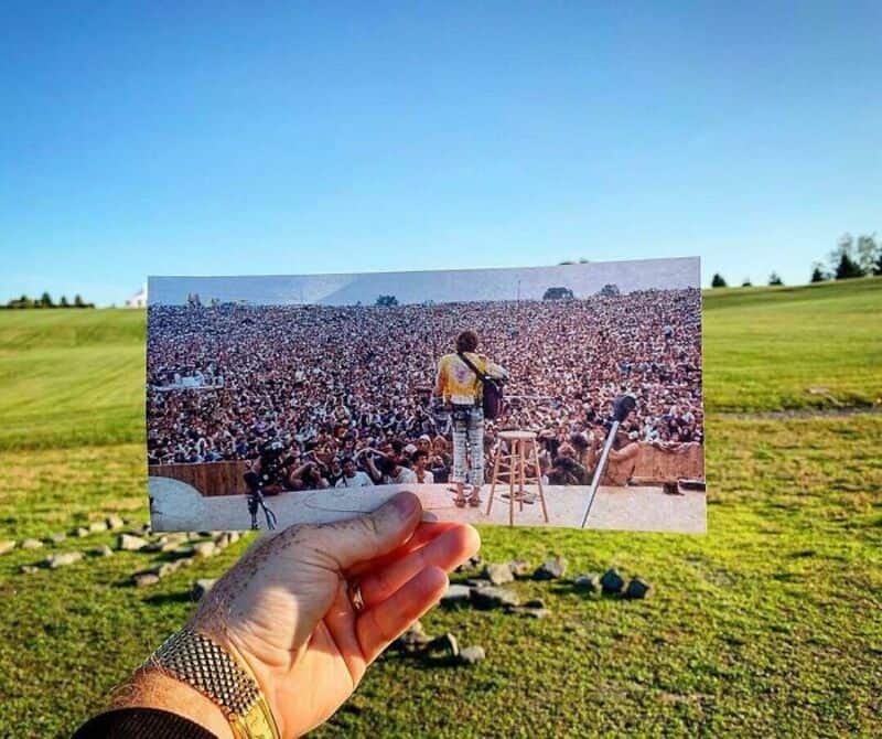 A hand holds a photo of a large crowd watching a performer on stage outdoors.