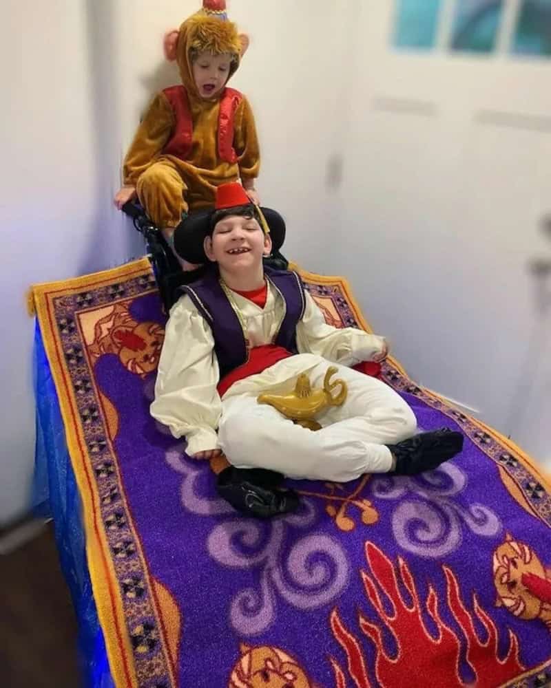 Two children in colorful costumes posed on a purple patterned blanket with a dragon design; one sits.