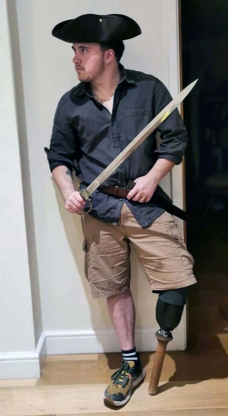 Man dressed as a cowboy holding a long sword, wearing a black hat, dark shirt, khaki shorts, and a l.