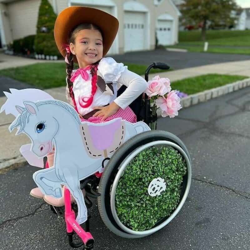 Young girl in a wheelchair dressed as a cowgirl with a pink saddle, hat, and decorative horse cutout.