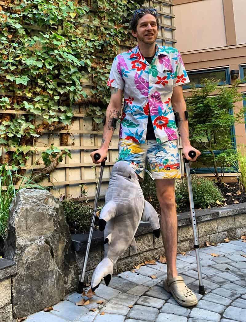 Man with crutches wearing a colorful floral shirt and shorts, holding a plush shark toy, outside on:.